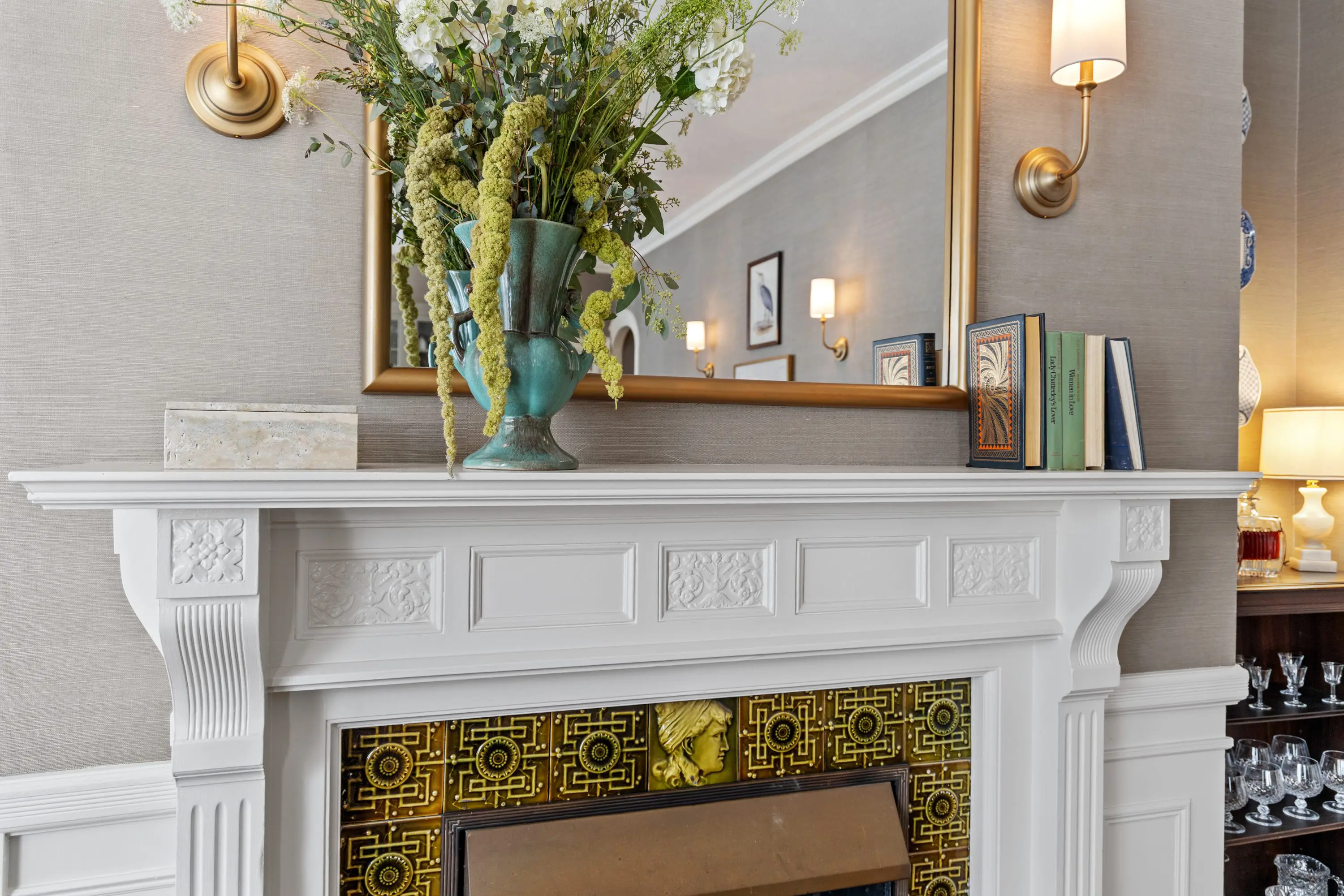 Elegant white fireplace with green floral arrangement and decorative mirror.