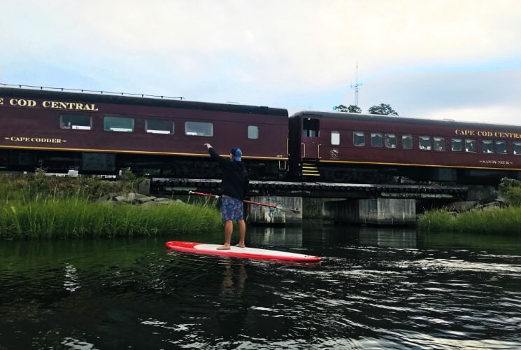 Person on stand up paddleboard in water waving to people on a moving train