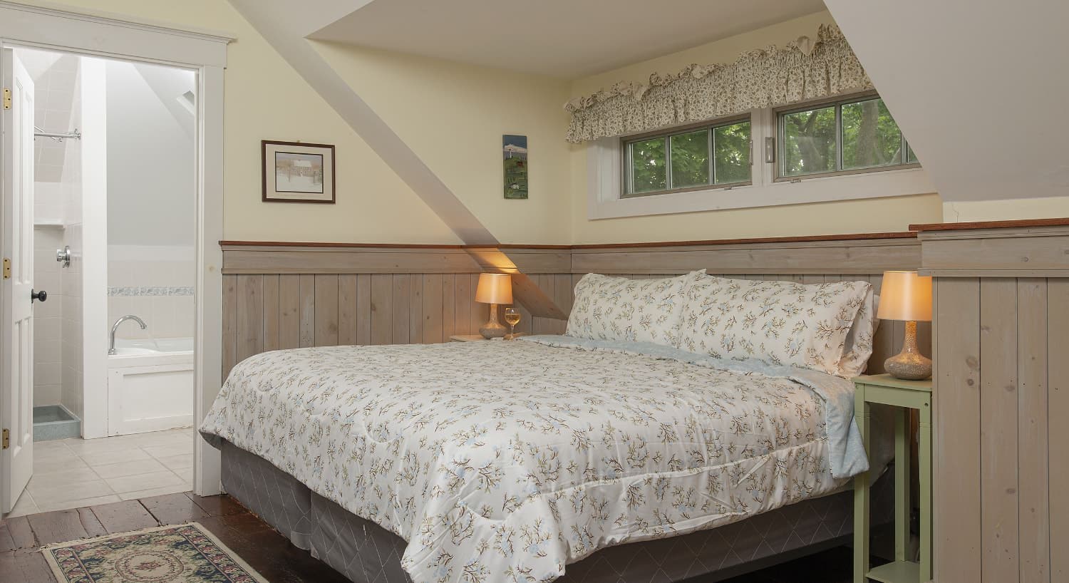 Bedroom with white walls, light wooden wainscoting, floral bedding, and view into bathroom