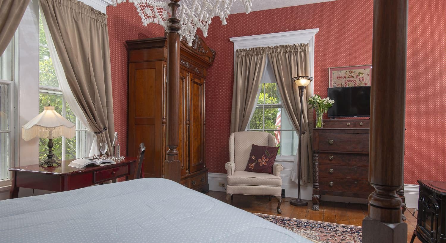 Bedroom with burgundy wallpaper, hardwood flooring, bed with light blue bedding, wooden desk, cream upholstered antique arm chair, and dark wooden hutch and dresser