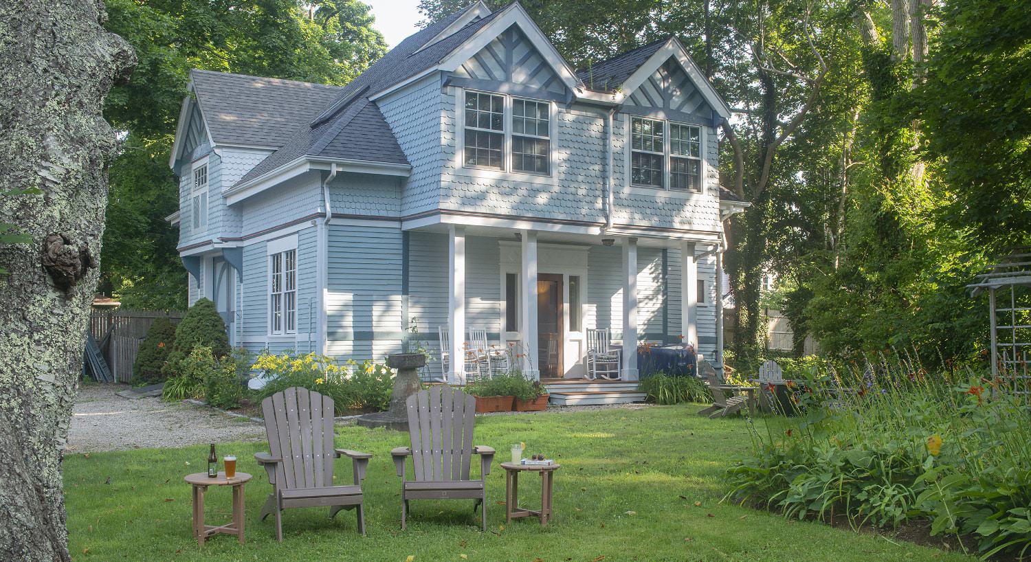 Exterior view of property painted light blue with white trim surrounded by large green trees and green yard with two Adirondack chairs