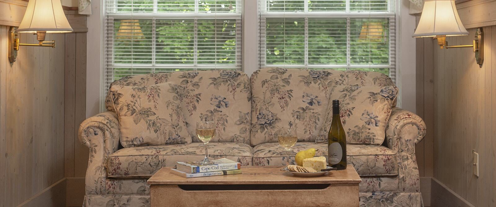 Floral upholstered loveseat and coffee table with Cape Cod books, wine filled glasses, and plate with cheese, crackers, and a pear