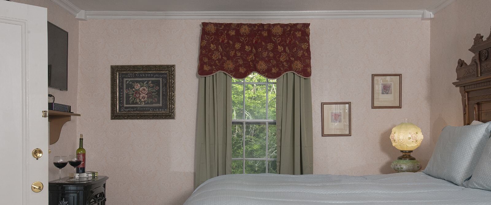 Bedroom with light pink brocade wallpaper, antique wooden headboard, light blue bedding, and window with a view of green trees