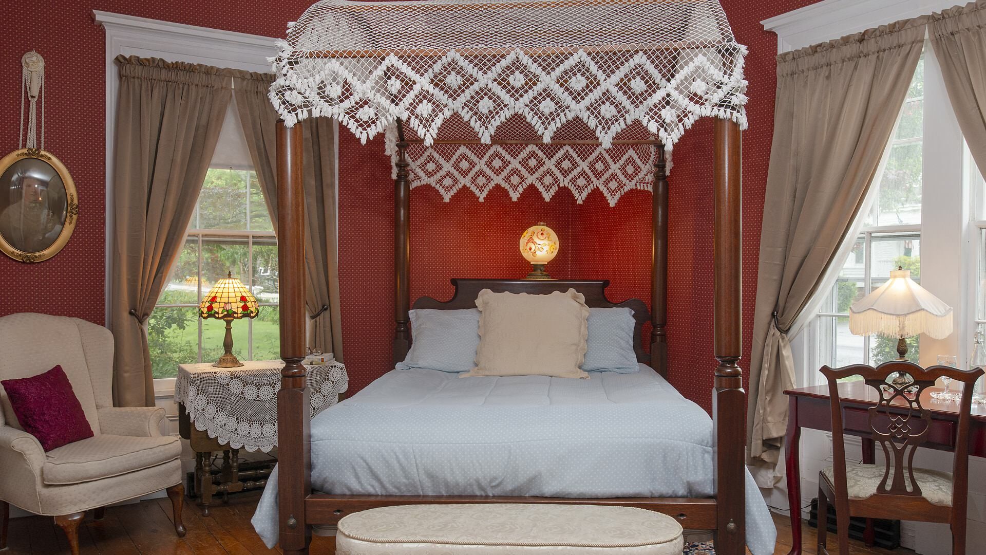 Bedroom with burgundy wallpaper, hardwood flooring, four-poster canopy dark wooden bed with light blue bedding, wooden desk, and cream upholstered antique arm chair