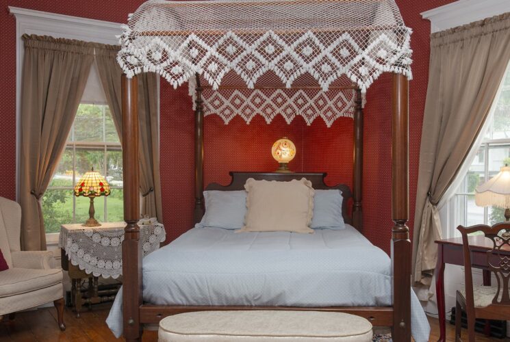 Bedroom with burgundy wallpaper, hardwood flooring, four-poster canopy dark wooden bed with light blue bedding, wooden desk, and cream upholstered antique arm chair