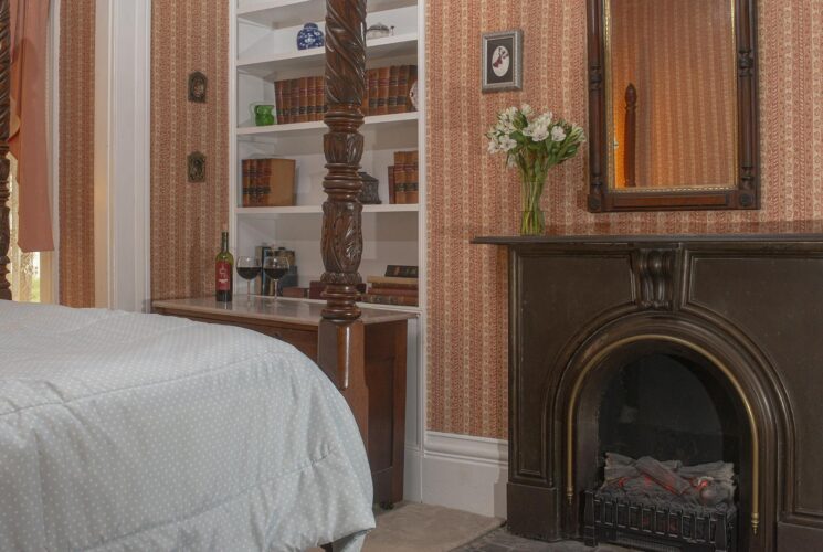 Bedroom with striped looking wallpaper, white trim, carpeting, four-poster wooden bed, and antique fireplace