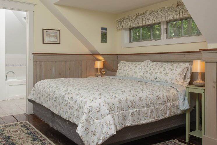 Bedroom with white walls, light wooden wainscoting, floral bedding, and view into bathroom